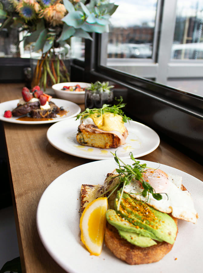 Mesa de brunch junto a la ventana con tostadas de palta y jugos